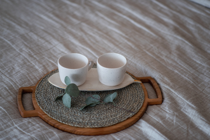 On the Linen bedspread lies a wooden round tray, on it are two empty ceramic cups, green eucalyptus